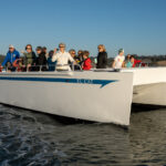 An excited group of passengers enjoying a delightful ride aboard the 'El Cat' during a privately chartered tour of Elkhorn Slough. The boat glides through serene waters, surrounded by lush coastal landscapes and diverse wildlife. Laughter and smiles fill the air as the passengers engage in lively conversations, captivated by the beauty of the slough's natural wonders. With binoculars in hand, some eagerly observe graceful seabirds soaring above, while others keep their eyes peeled for curious otters playing in the water. The warm sunlight bathes the scene in a golden glow, creating a picture-perfect moment of relaxation, exploration, and camaraderie.