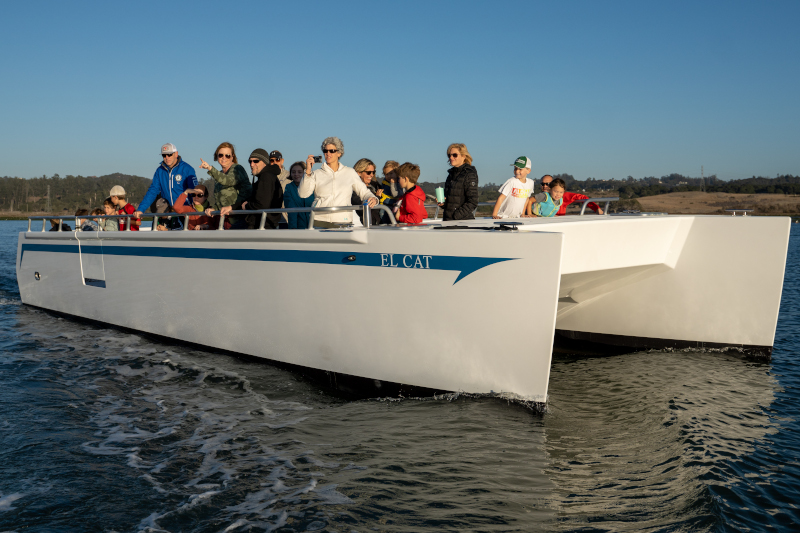 An excited group of passengers enjoying a delightful ride aboard the 'El Cat' during a privately chartered tour of Elkhorn Slough. The boat glides through serene waters, surrounded by lush coastal landscapes and diverse wildlife. Laughter and smiles fill the air as the passengers engage in lively conversations, captivated by the beauty of the slough's natural wonders. With binoculars in hand, some eagerly observe graceful seabirds soaring above, while others keep their eyes peeled for curious otters playing in the water. The warm sunlight bathes the scene in a golden glow, creating a picture-perfect moment of relaxation, exploration, and camaraderie.